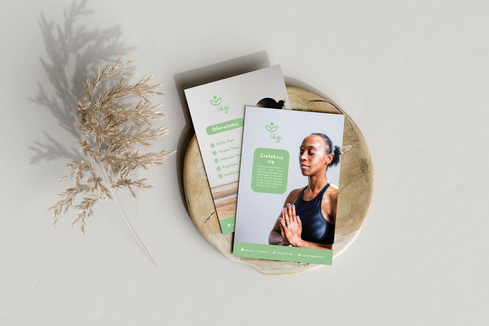 Dwie wizytówki studia jogi 'lõgi' na drewnianej podstawce, z dekoracyjną gałązką trawy w tle. Jedna wizytówka prezentuje ofertę zajęć, druga przedstawia kobietę w pozycji medytacyjnej.