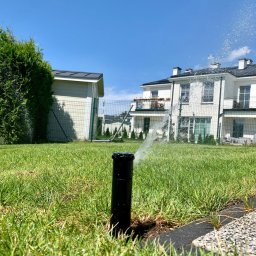 Velto - Niski kąt ujęcia na działający, czarny zraszacz trawnikowy, z widocznymi kroplami wody w powietrzu, na tle zielonego trawnika, ogrodzenia i białego domu z balkonem.