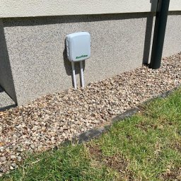 Velto - Szary kontroler nawadniania Rain Bird zamontowany na elewacji budynku, otoczony ozdobnymi kamieniami i trawnikiem.