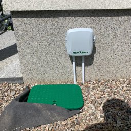 Velto - Szara skrzynka sterująca systemem nawadniania Rain Bird przymocowana do elewacji budynku, zielona pokrywa zaworu w ziemi, otoczenie wysypane kamykami.