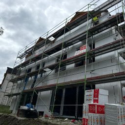 Velto - Budynek wielorodzinny w trakcie ocieplania, widoczne rusztowania, styropianowe płyty ociepleniowe i pracownik w kamizelce odblaskowej na rusztowaniu. Budynek wykonany z białych bloczków.