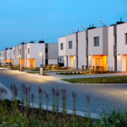 Nowoczesne osiedle domów szeregowych z białymi elewacjami i pomarańczowymi wnękami wejściowymi, oświetlone lampami ulicznymi o zmierzchu, z widoczną zielenią i chodnikiem z kostki brukowej.