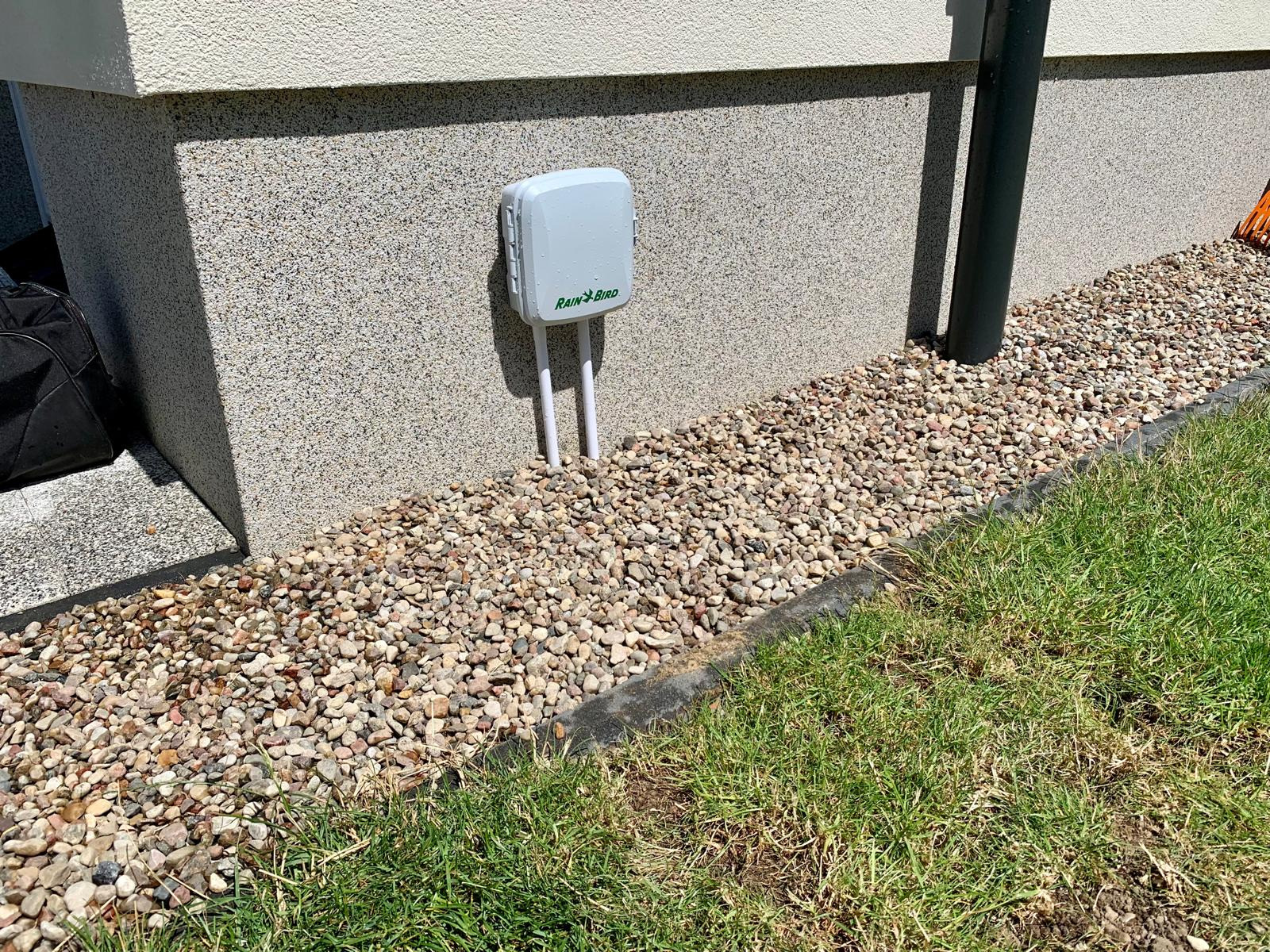 Szary kontroler nawadniania Rain Bird zamontowany na elewacji budynku, otoczony ozdobnymi kamieniami i trawnikiem.