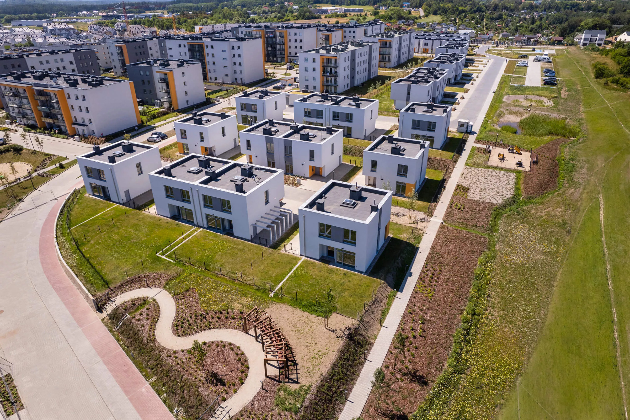 Nowoczesne osiedle domów jednorodzinnych w Gdyni, widok z lotu ptaka ukazuje szereg identycznych, białych budynków z szarymi akcentami, zadbane trawniki i ścieżki spacerowe, a w tle rozległe tereny...