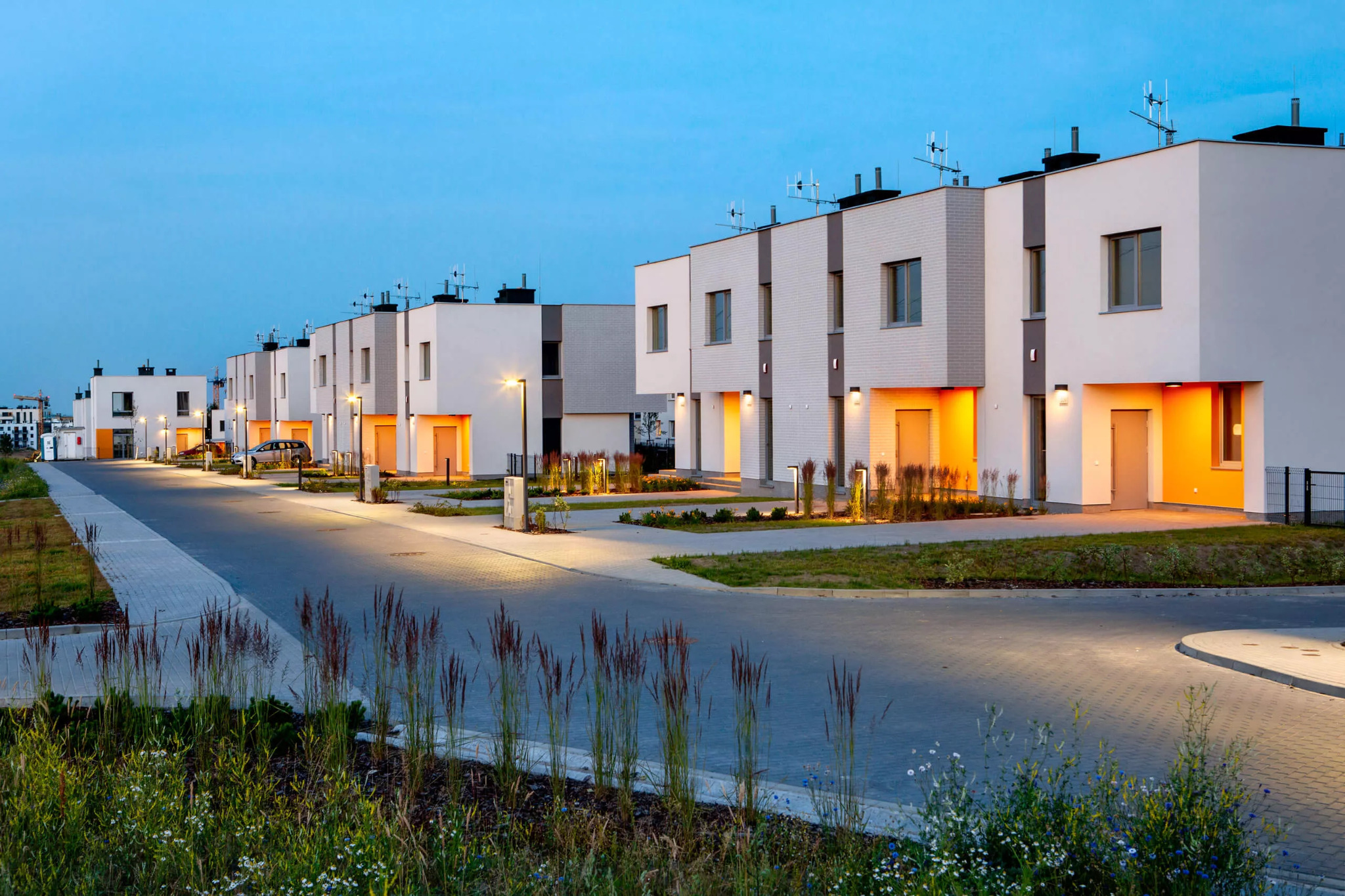 Nowoczesne osiedle domów szeregowych z białymi elewacjami i pomarańczowymi wnękami wejściowymi, oświetlone lampami ulicznymi o zmierzchu, z widoczną zielenią i chodnikiem z kostki brukowej.