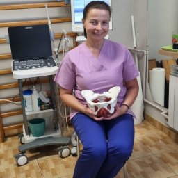 GABINET FIZJOTERAPII GABRIELA RATAJSKA - Uśmiechnięta kobieta w uniformie medycznym siedzi i trzyma model miednicy w gabinecie z drabinką rehabilitacyjną i aparaturą USG w tle.
