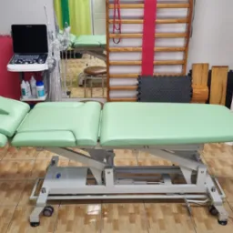 Jasnozielony stół do terapii z regulacją wysokości, obok aparat USG, drabinki gimnastyczne i wyposażenie gabinetu.