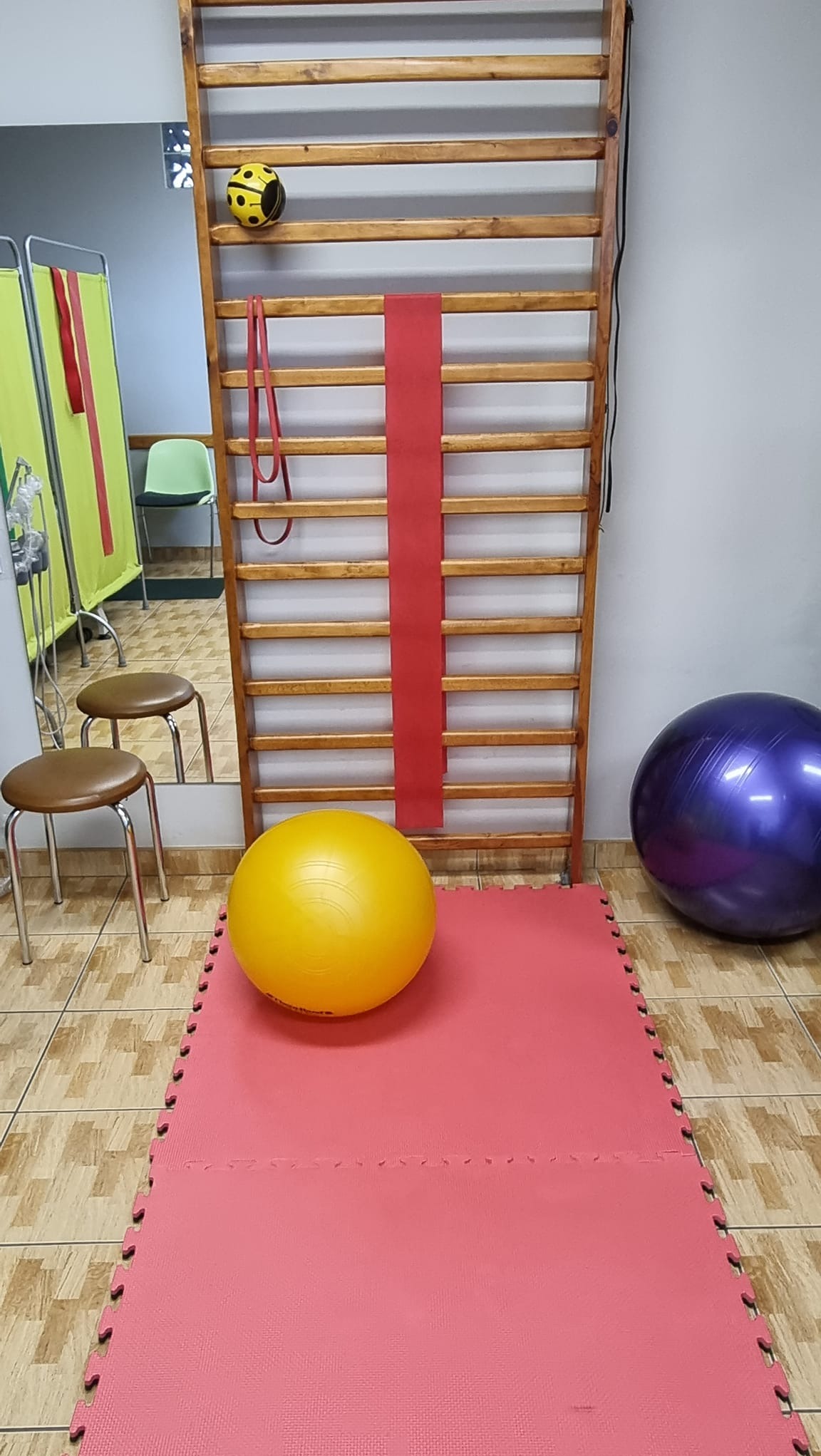 Sala ćwiczeń z drabinką gimnastyczną, żółtą piłką, fioletową piłką do ćwiczeń, czerwoną matą i gumami do ćwiczeń.