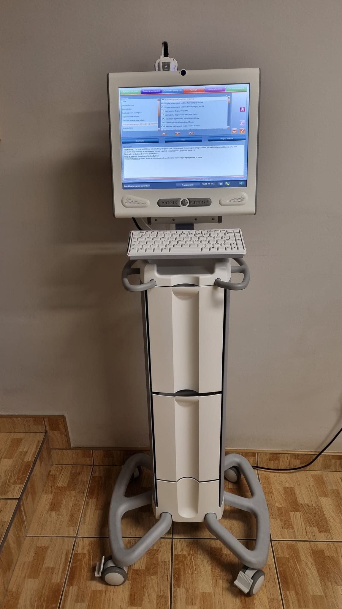 Mobilne urządzenie diagnostyczne Phenix Liberty z ekranem dotykowym, klawiaturą i oprogramowaniem terapeutycznym, na kółkach, w gabinecie z brązowymi płytkami podłogowymi.