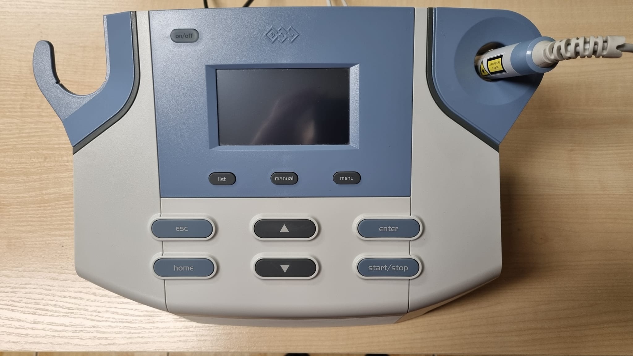 Urządzenie do laseroterapii z ekranem LCD i przyciskami funkcyjnymi, widoczna końcówka lasera z ostrzeżeniem, na drewnianym blacie.