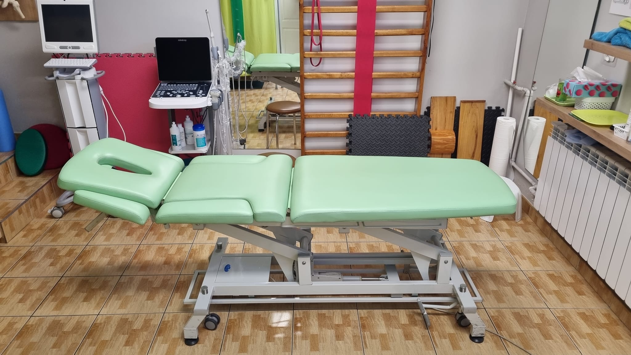 Jasnozielony stół do terapii z regulacją wysokości, obok aparat USG, drabinki gimnastyczne i wyposażenie gabinetu.