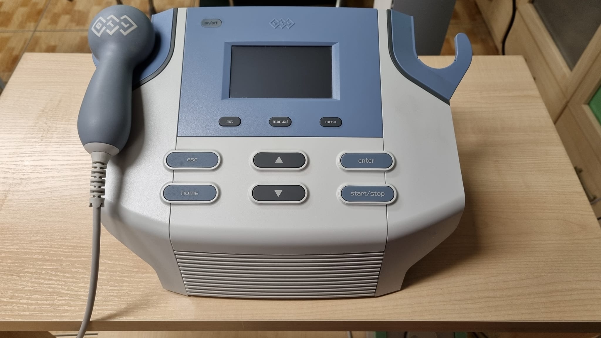 Urządzenie do fizykoterapii z aplikatorem ultradźwiękowym, ekranem i przyciskami funkcyjnymi, umieszczone na drewnianym stole.