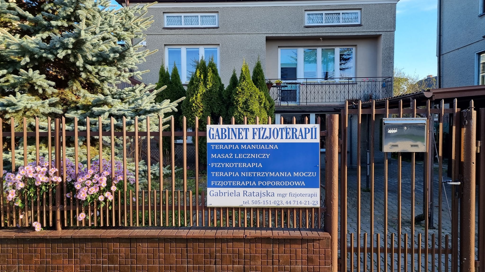 Tablica informacyjna Gabinetu Fizjoterapii z ofertą terapii manualnej, masażu leczniczego, fizykoterapii i terapii nietrzymania moczu, umieszczona na brązowym ogrodzeniu przed budynkiem.