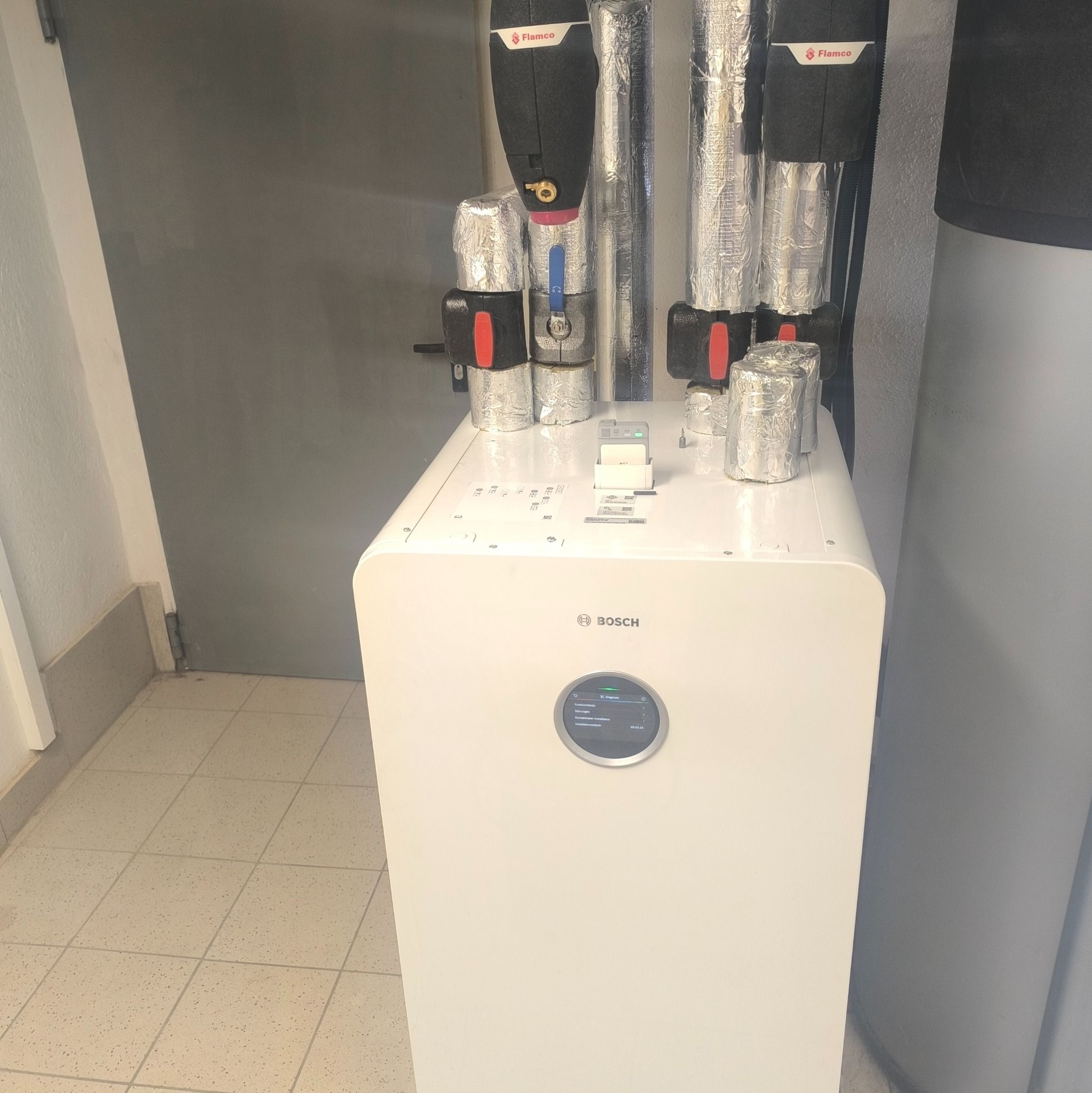 Nowoczesny, biały piec gazowy Bosch z podłączonymi rurami i zaworami, zabezpieczonymi folią aluminiową. Widoczne elementy instalacji grzewczej w pomieszczeniu.