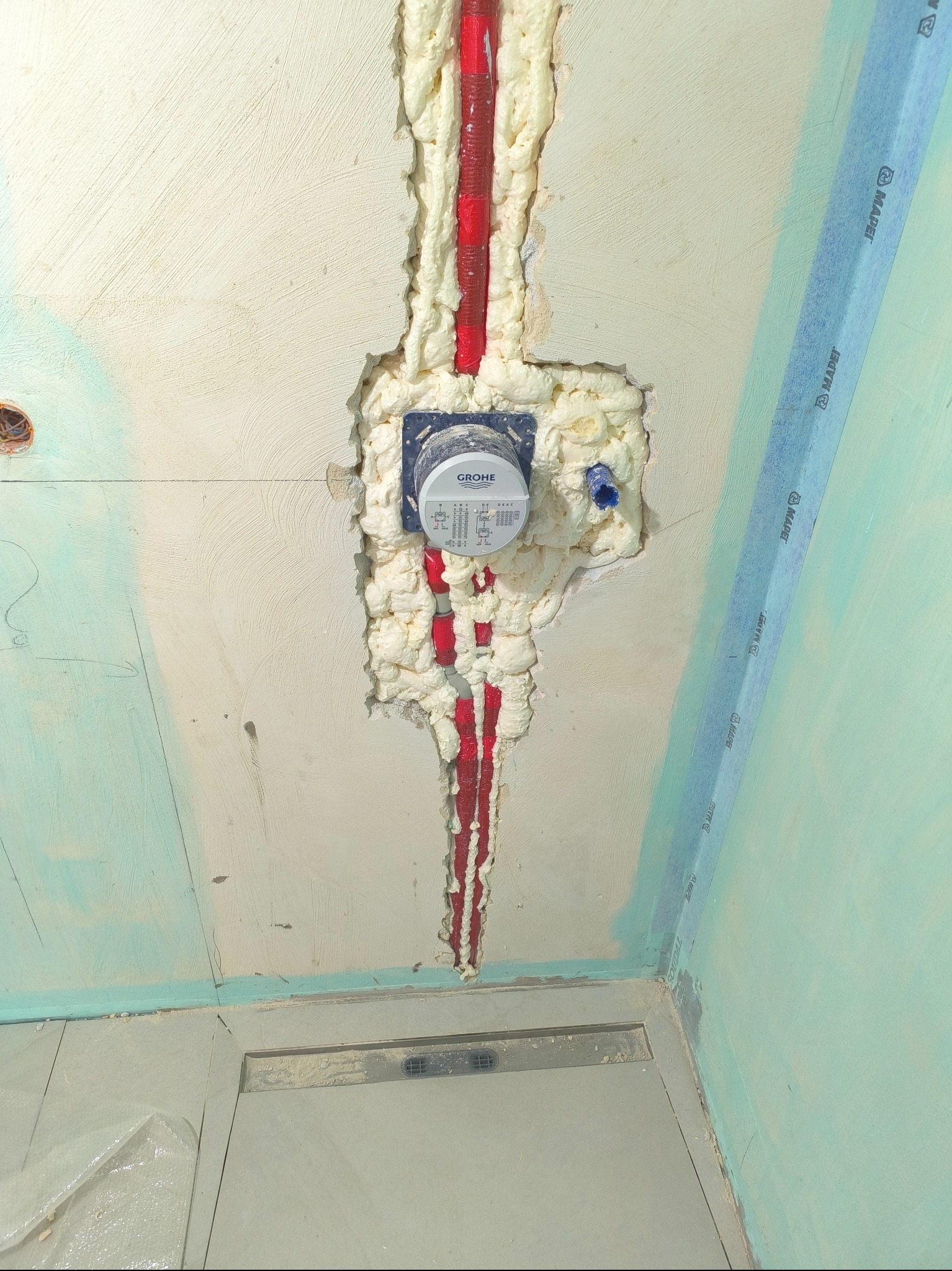 Ściana z zamontowanym, zaizolowanym pianką i rurami wodomierzem marki Grohe. Widoczne ślady po taśmie malarskiej i fragment odpływu liniowego w podłodze.