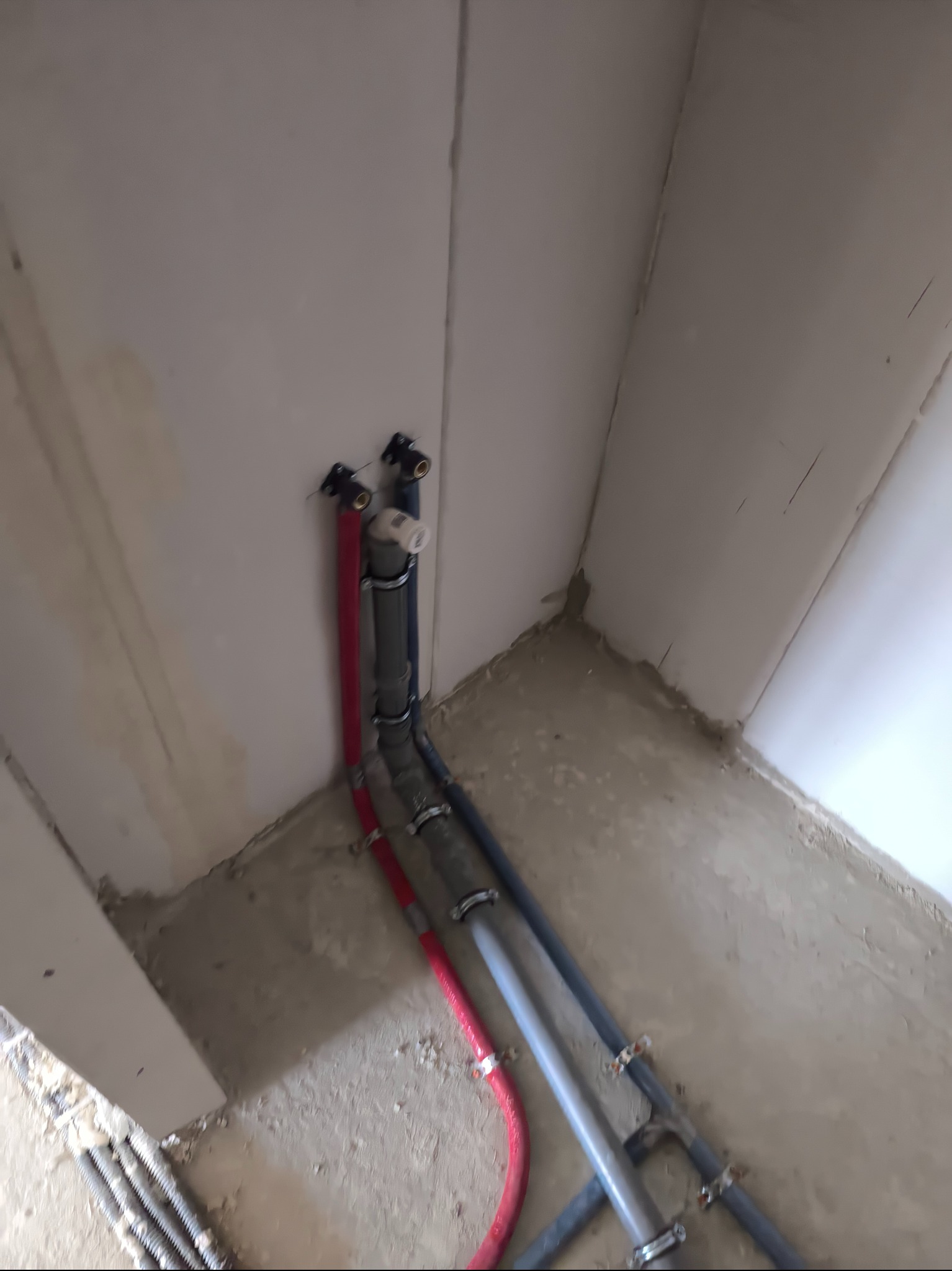 Ukończona instalacja wodno-kanalizacyjna w rogu pomieszczenia. Widoczne rury z tworzywa sztucznego w kolorach szarym i czerwonym, przymocowane do ściany za pomocą metalowych uchwytów.