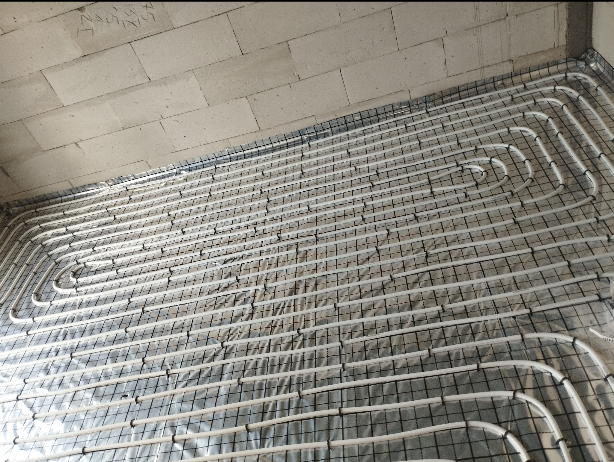 Białe rury ogrzewania podłogowego ułożone na srebrnej folii, przymocowane do czarnej siatki, widok z góry, surowy mur z betonu komórkowego w tle.