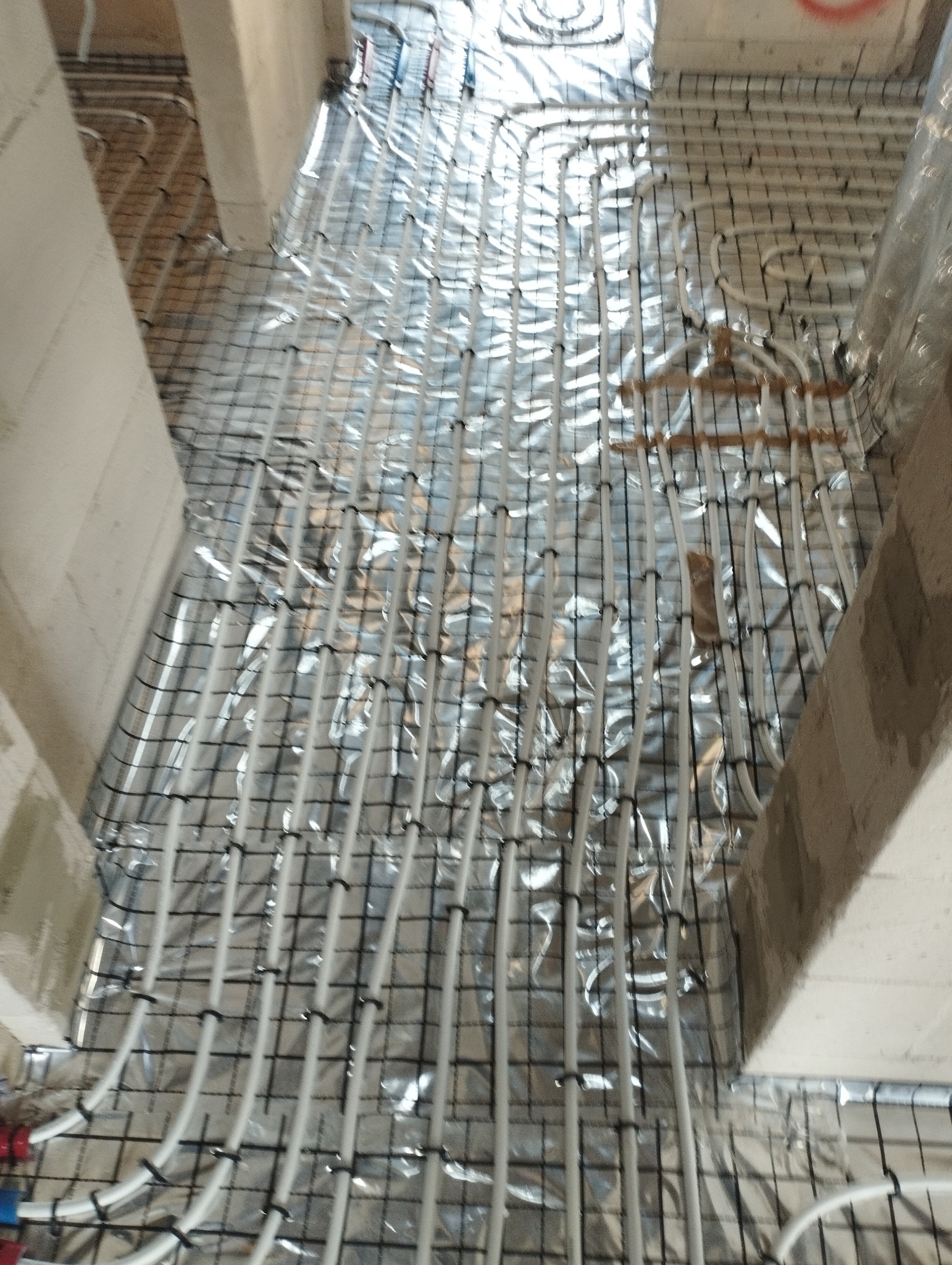 Białe rury ogrzewania podłogowego ułożone na folii izolacyjnej i siatce zbrojeniowej, widok z góry, surowy stan budynku, przygotowanie do wylewki.