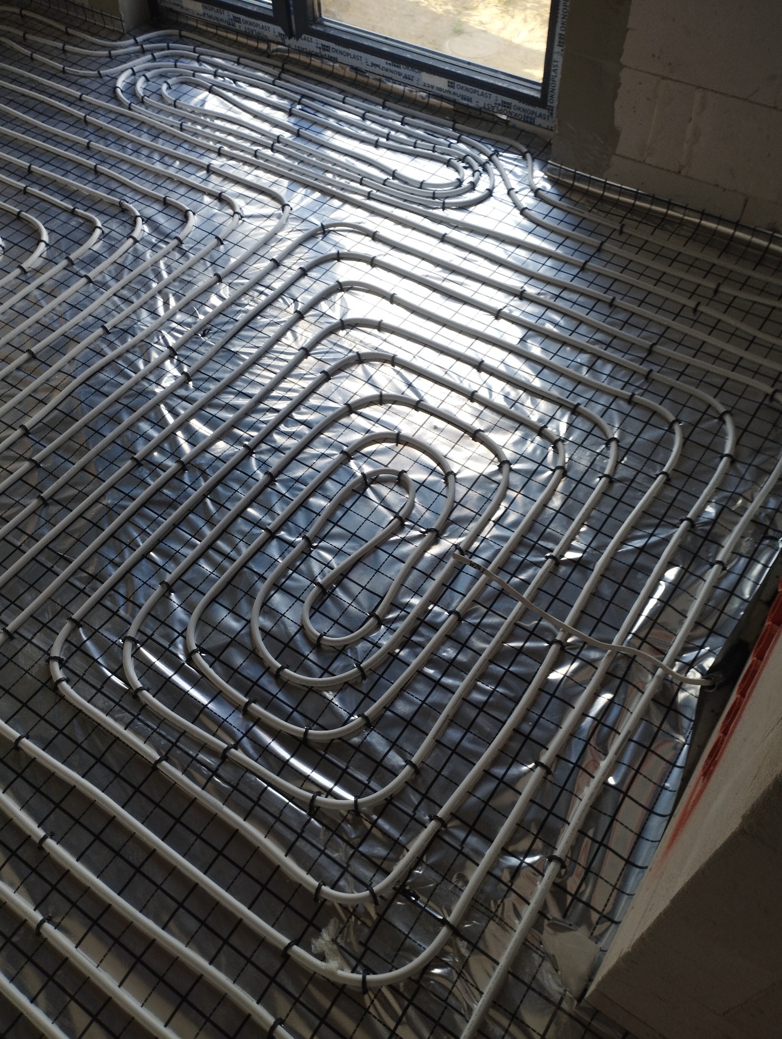 Białe rury ogrzewania podłogowego ułożone na folii izolacyjnej z czarną siatką, widoczne okno i fragment ściany. Układ spiralny rur, etap instalacji przed wylewką.