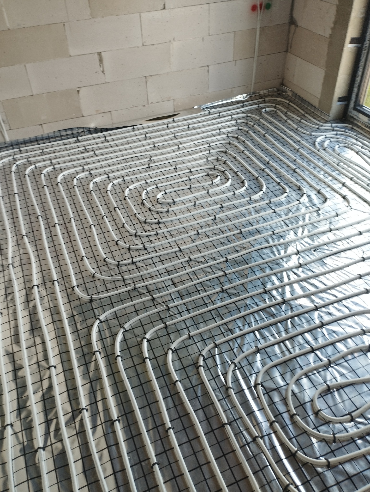 Białe rury ogrzewania podłogowego ułożone spiralnie na srebrnej folii izolacyjnej, przymocowane czarnymi uchwytami do siatki. Ściany z białych bloczków.