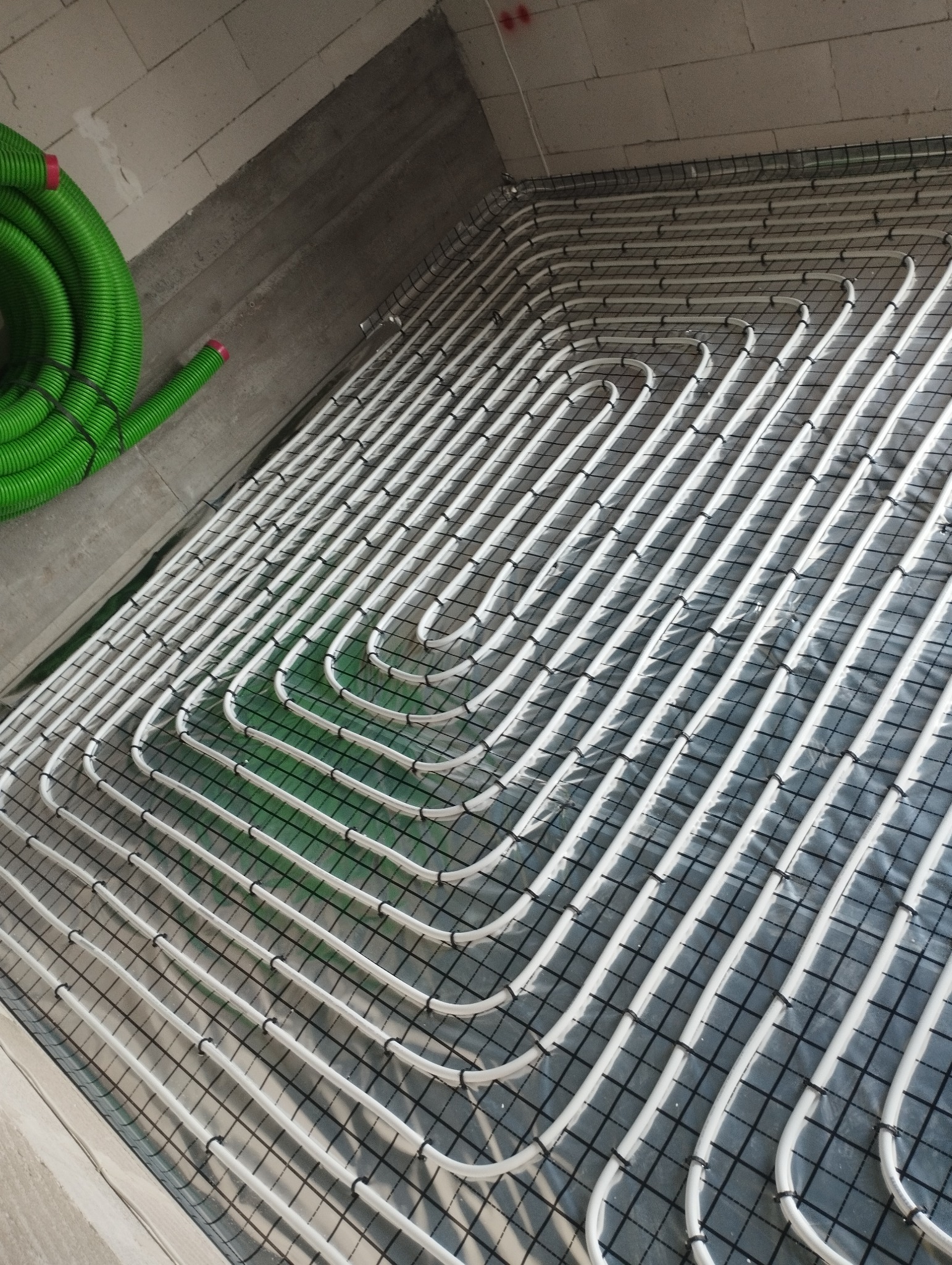 Białe rury ogrzewania podłogowego ułożone w pętle na czarnej siatce montażowej, z zielonym przewodem wentylacyjnym w rogu. Surowy beton na ścianach i suficie.
