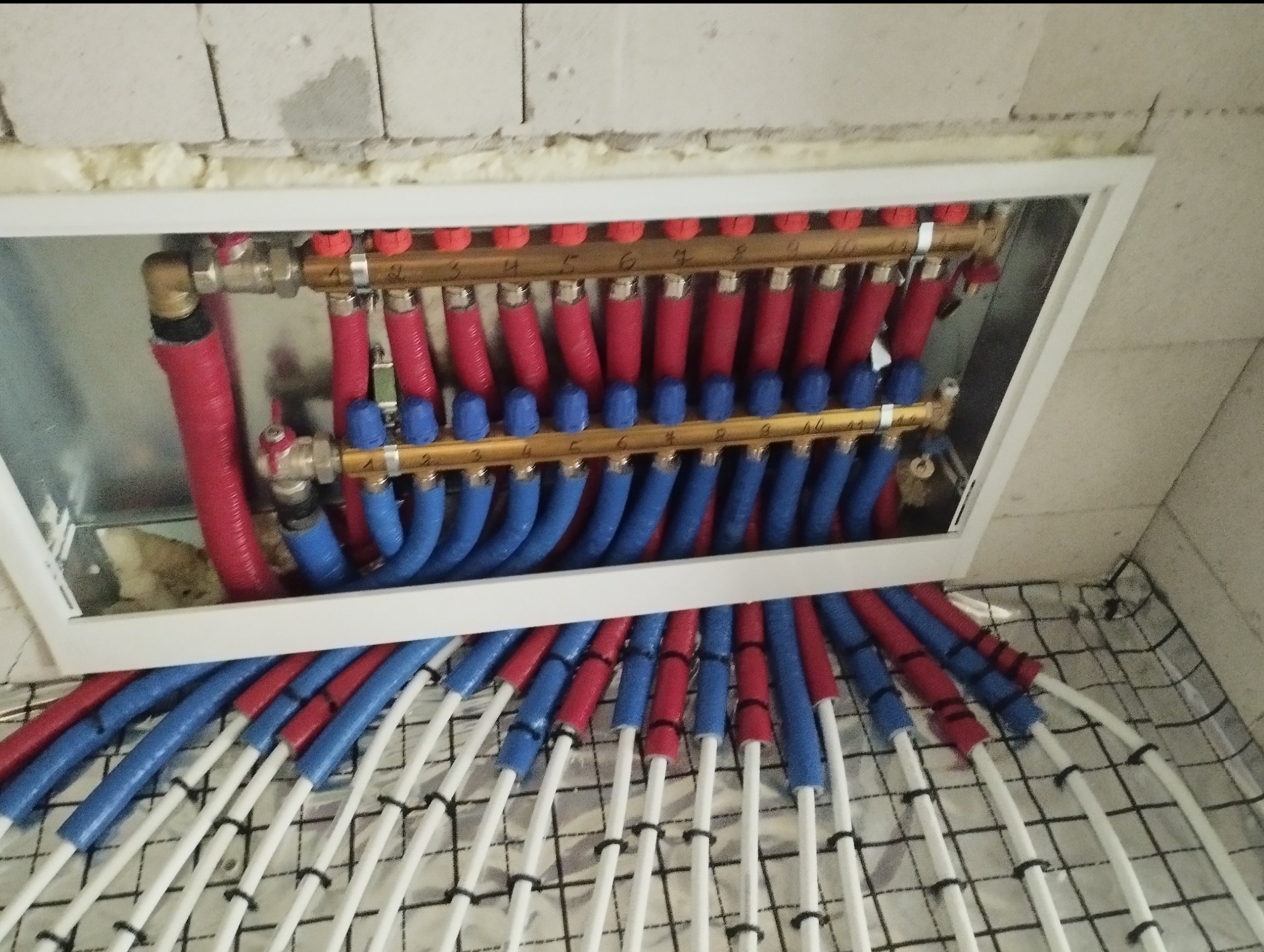 Rozdzielacz ogrzewania podłogowego w ramie, z czerwonymi i niebieskimi rurami, zamontowany na siatce. Widoczne elementy instalacji i fragment ściany.
