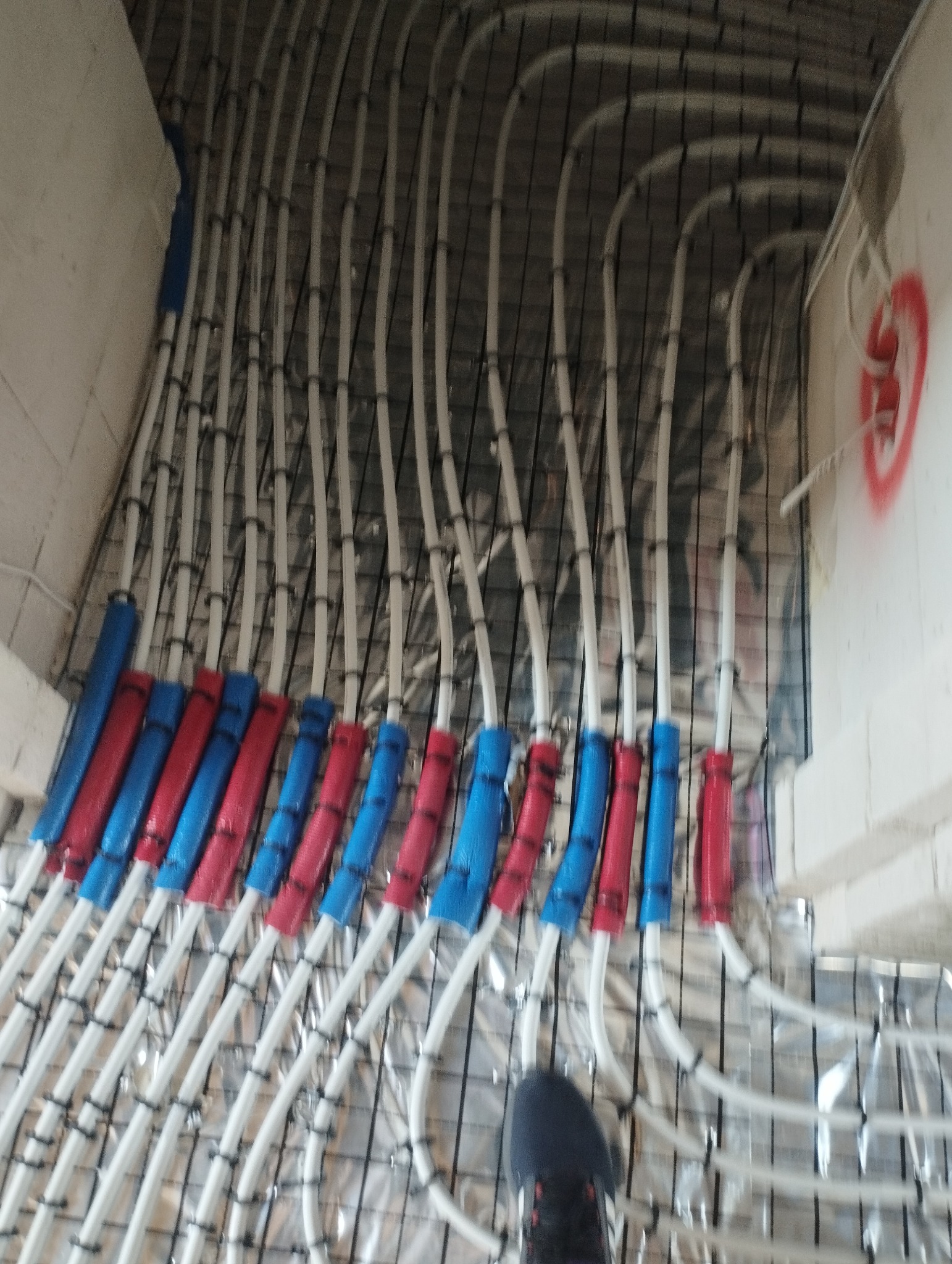 Białe rury ogrzewania podłogowego, zabezpieczone kolorowymi otulinami (czerwone i niebieskie), ułożone na metalowej siatce z folią. Widoczny fragment buta.