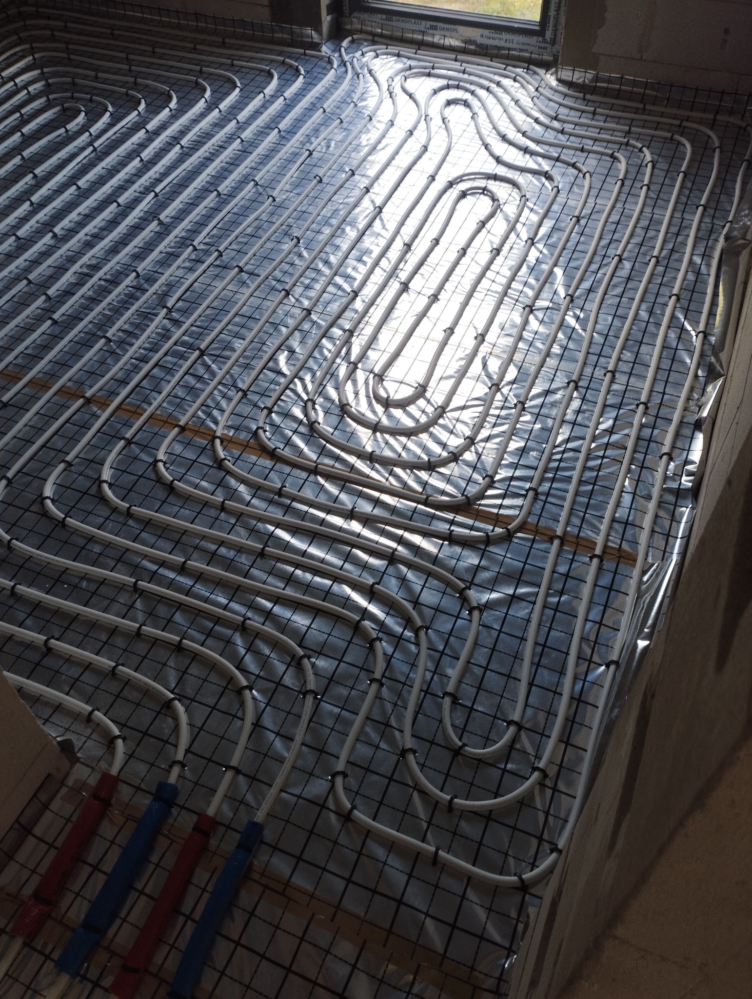 Białe rury ogrzewania podłogowego ułożone na folii z siatką, widoczne niebieskie i czerwone końcówki rur, okno w tle. Instalacja w trakcie realizacji.