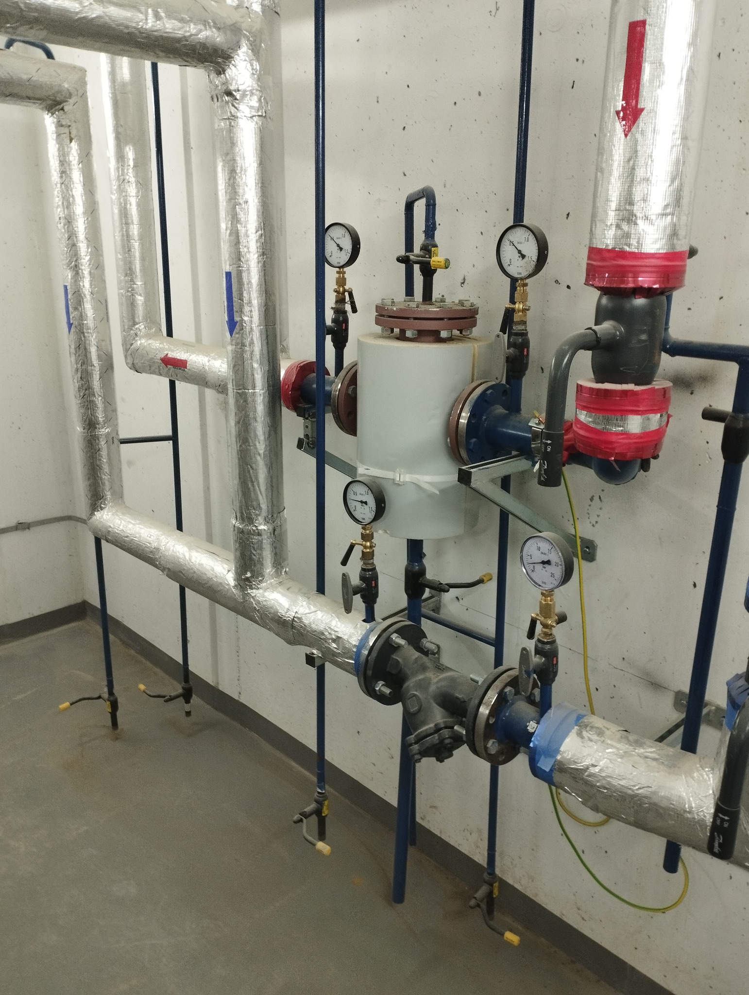 Złożona instalacja rur z izolacją aluminiową, manometry i zawory na tle szarej ściany i podłogi. Widoczne niebieskie podpory i strzałki wskazujące kierunek przepływu.