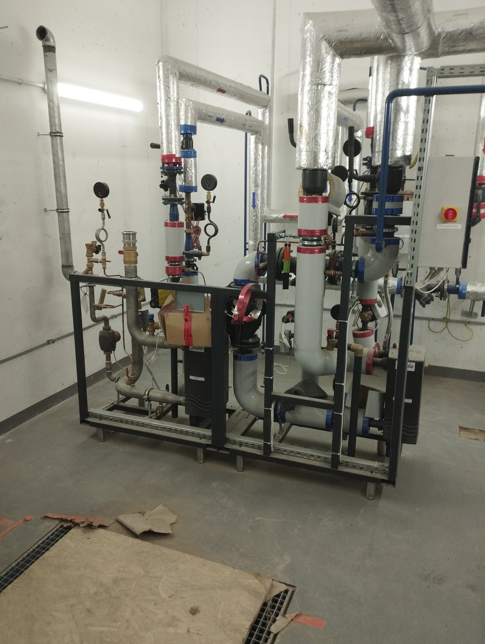 Złożona instalacja grzewcza z rurami izolowanymi folią aluminiową, zaworami i manometrami, umieszczona na metalowej ramie w pomieszczeniu technicznym.