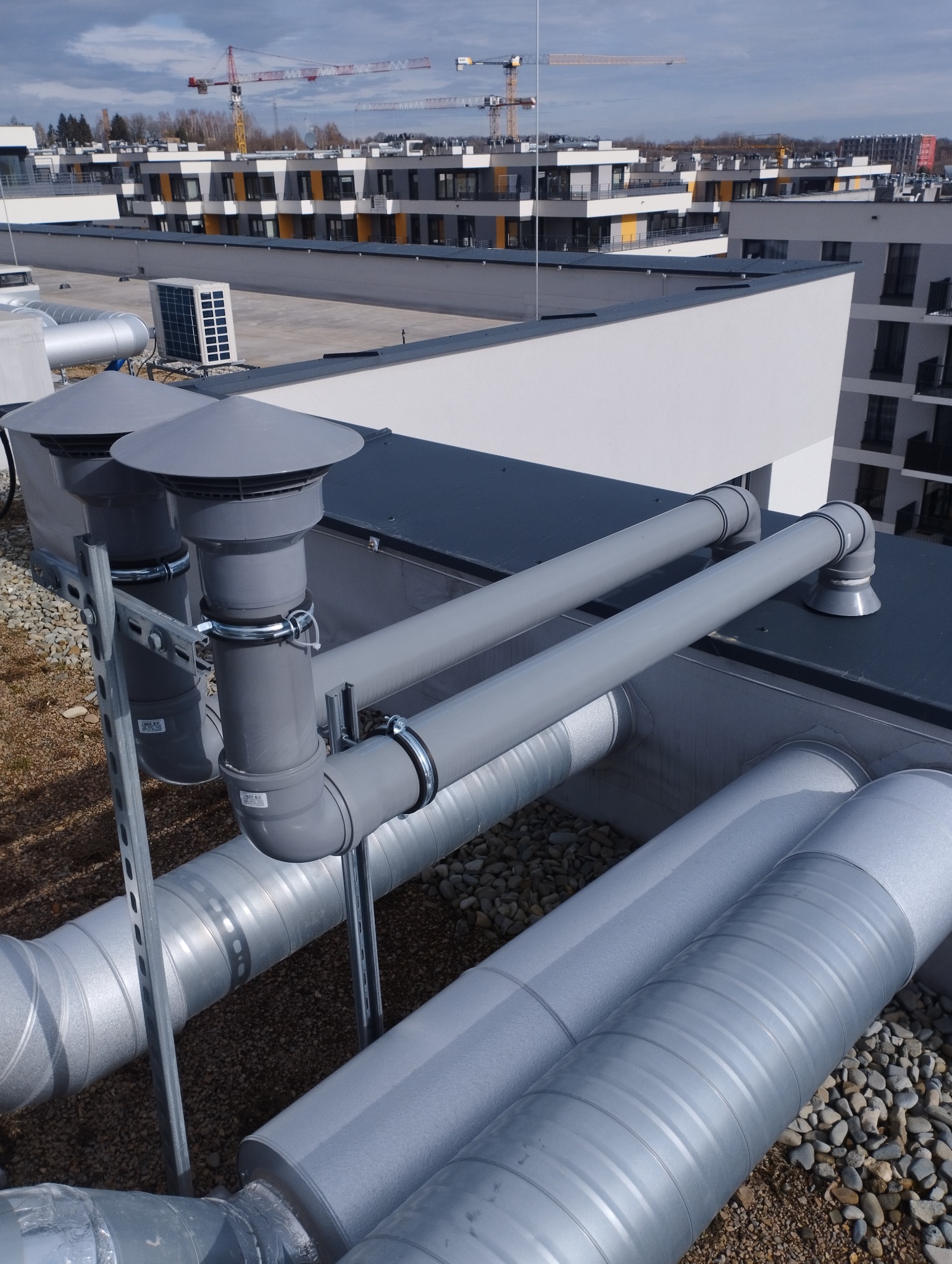 Szare rury wentylacyjne na dachu budynku z widokiem na nowe budownictwo i żurawie budowlane w tle. Instalacja na żwirowym podłożu, z elementami mocującymi.