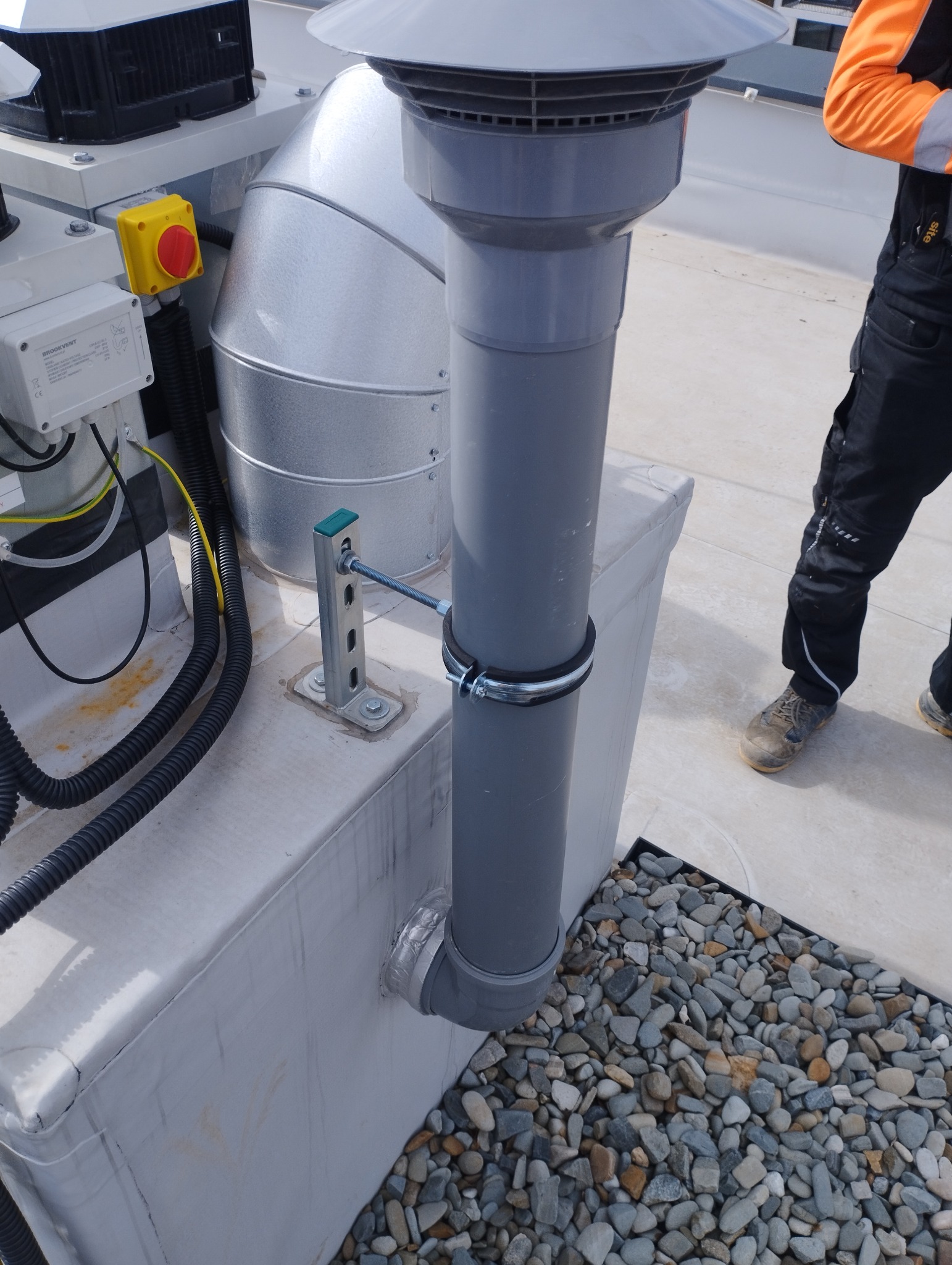 Szara rura wentylacyjna z metalowym kołnierzem i obejmą, zamocowana do białej obudowy na dachu, z widocznymi kablami i fragmentem osoby w roboczym stroju.