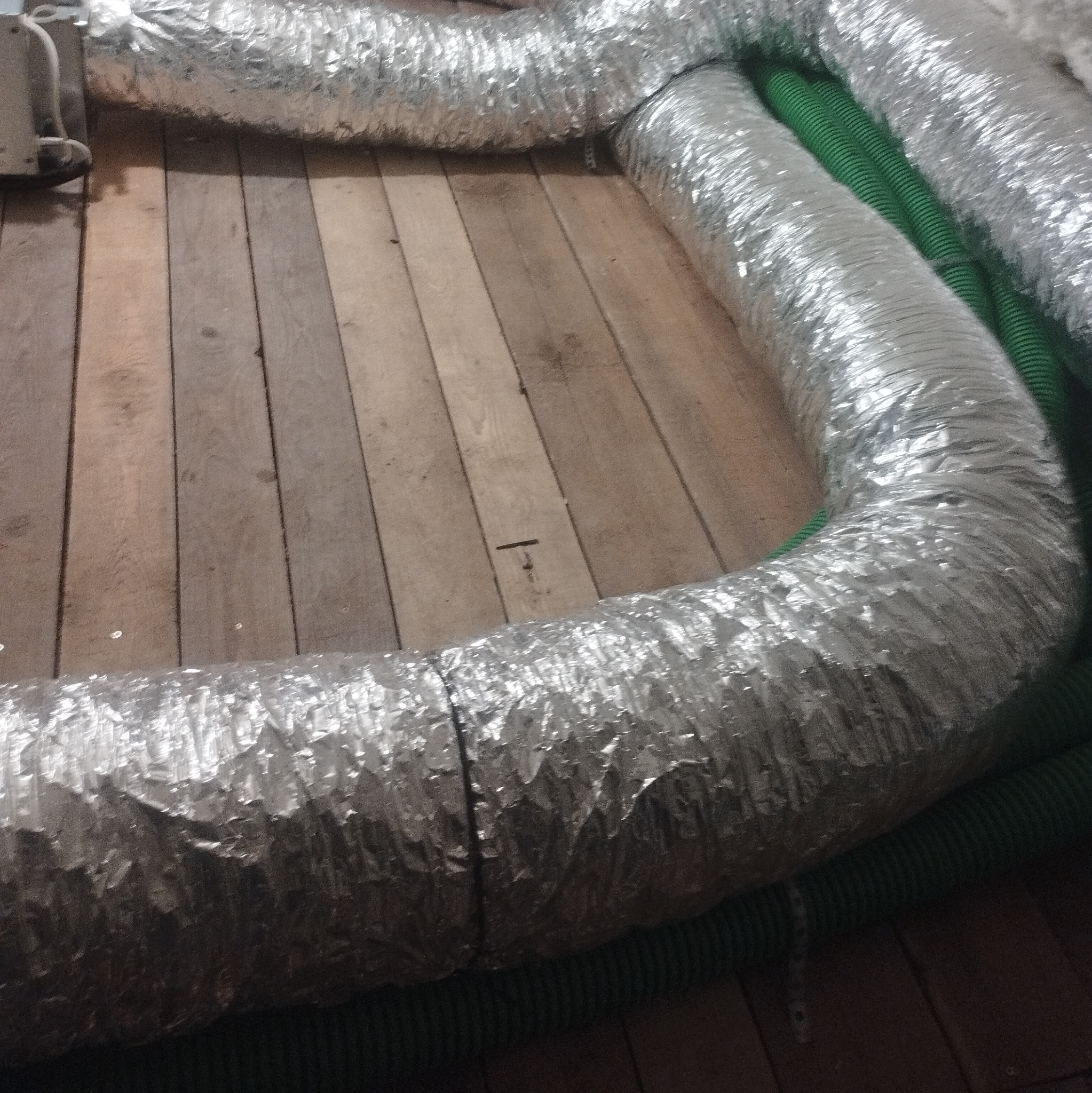 Elastyczne, aluminiowe przewody wentylacyjne ułożone na drewnianej podłodze, częściowo zakryte zielonymi rurami karbowanymi. Widoczne łączenia przewodów.