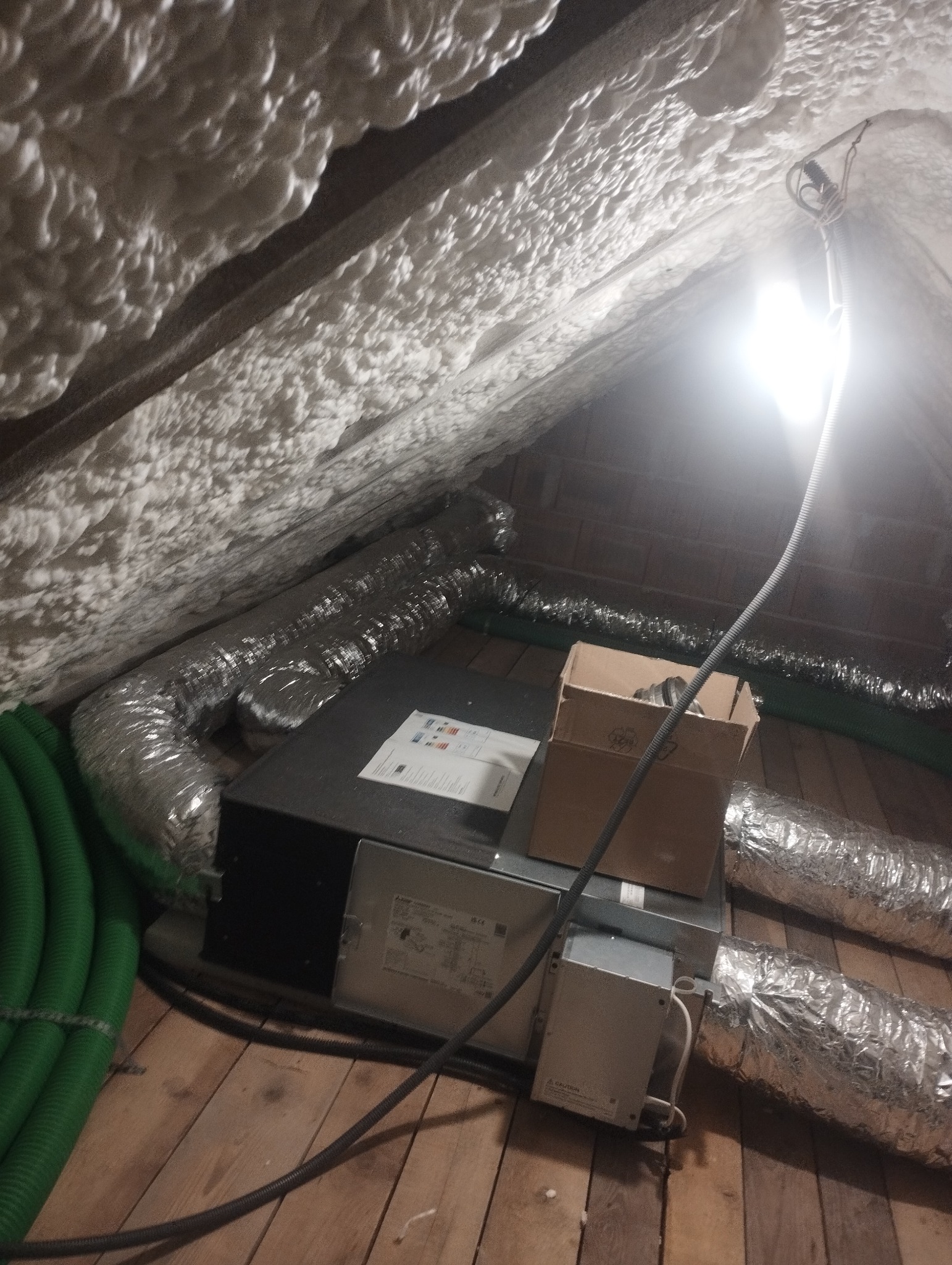Instalacja wentylacyjna na poddaszu z izolacją piankową, widoczne przewody wentylacyjne, skrzynka rozdzielcza i lampa oświetlająca przestrzeń roboczą.