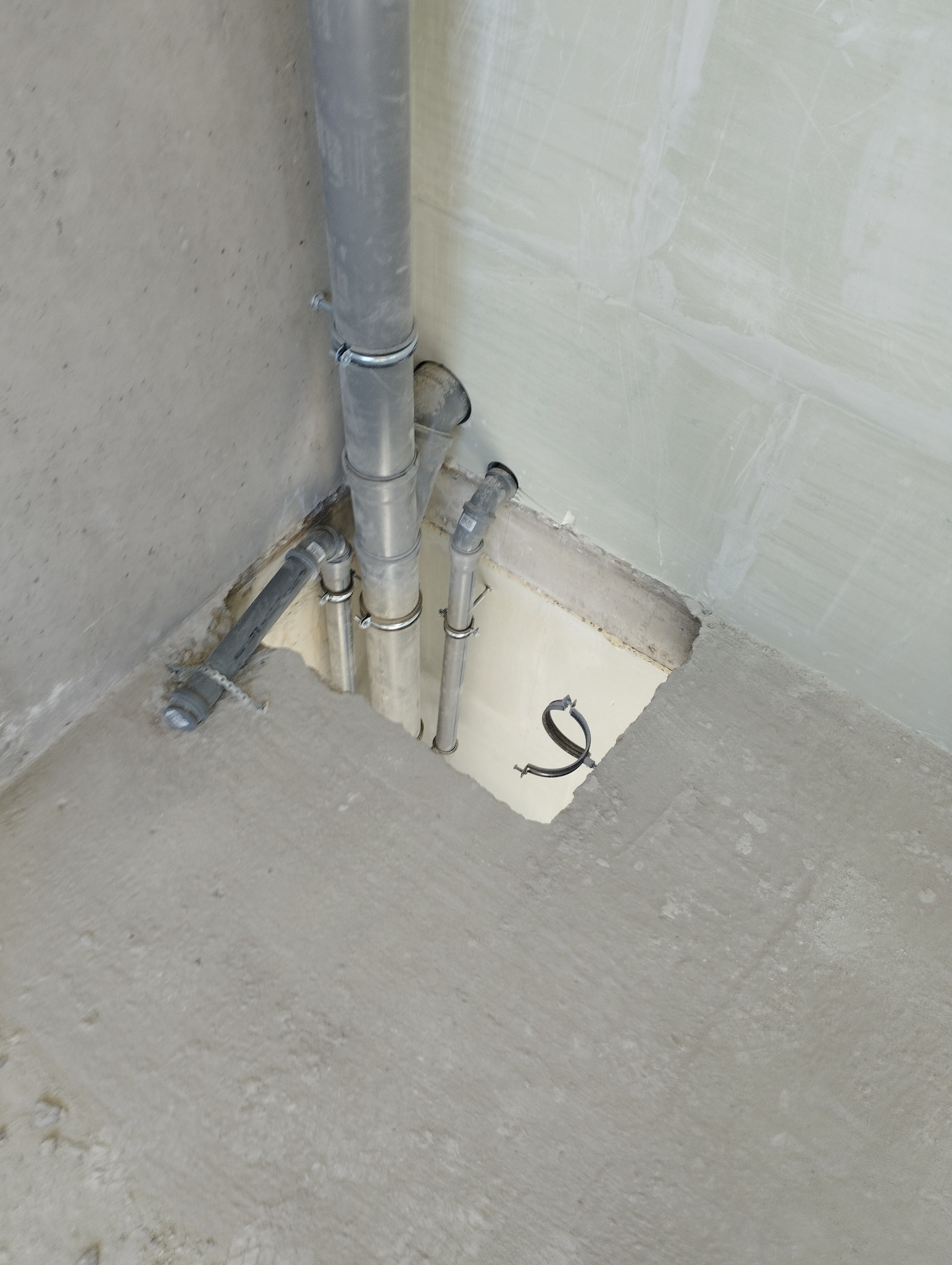 Szare rury kanalizacyjne z metalowymi obejmami, wystające z dziury w betonowej podłodze przy ścianie. Widoczne fragmenty instalacji i surowy stan wykończenia.