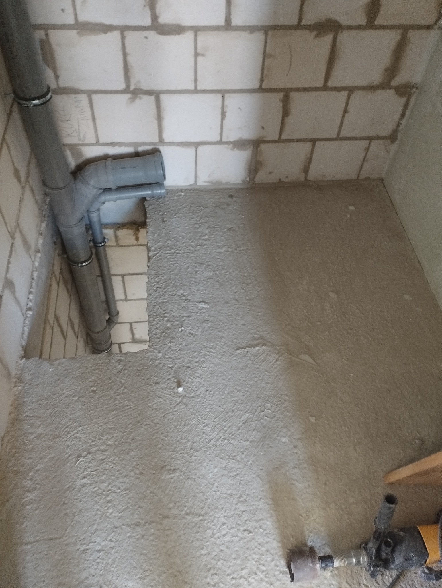 Szare rury kanalizacyjne z obejmami, wmurowane w ścianę z białych bloczków, z widocznym fragmentem podłogi w trakcie wylewki. W tle ściana.