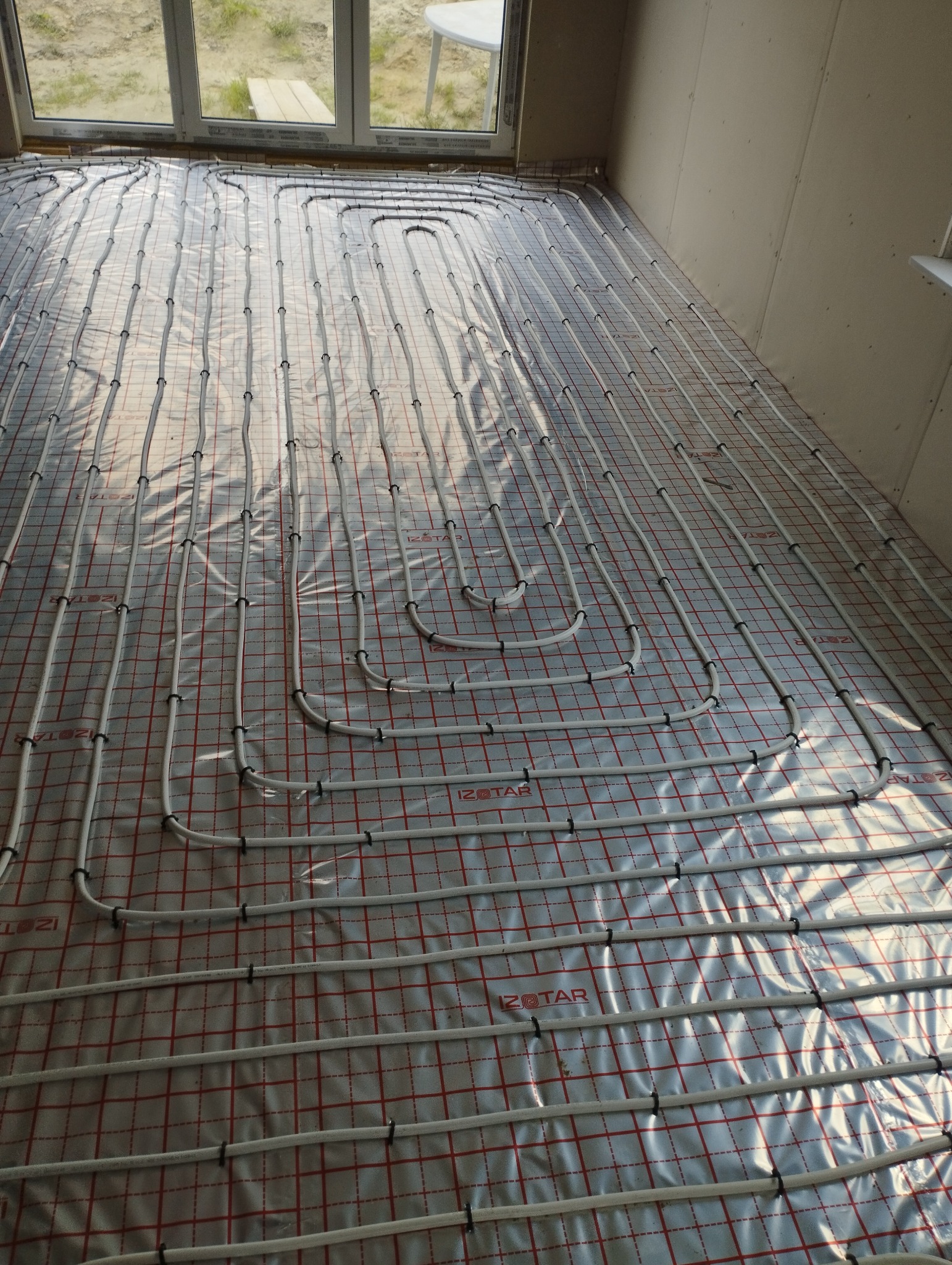 Białe rury ogrzewania podłogowego ułożone na folii z czerwoną siatką w jasnym pomieszczeniu z oknem. Widok z góry, instalacja przed wylaniem posadzki.