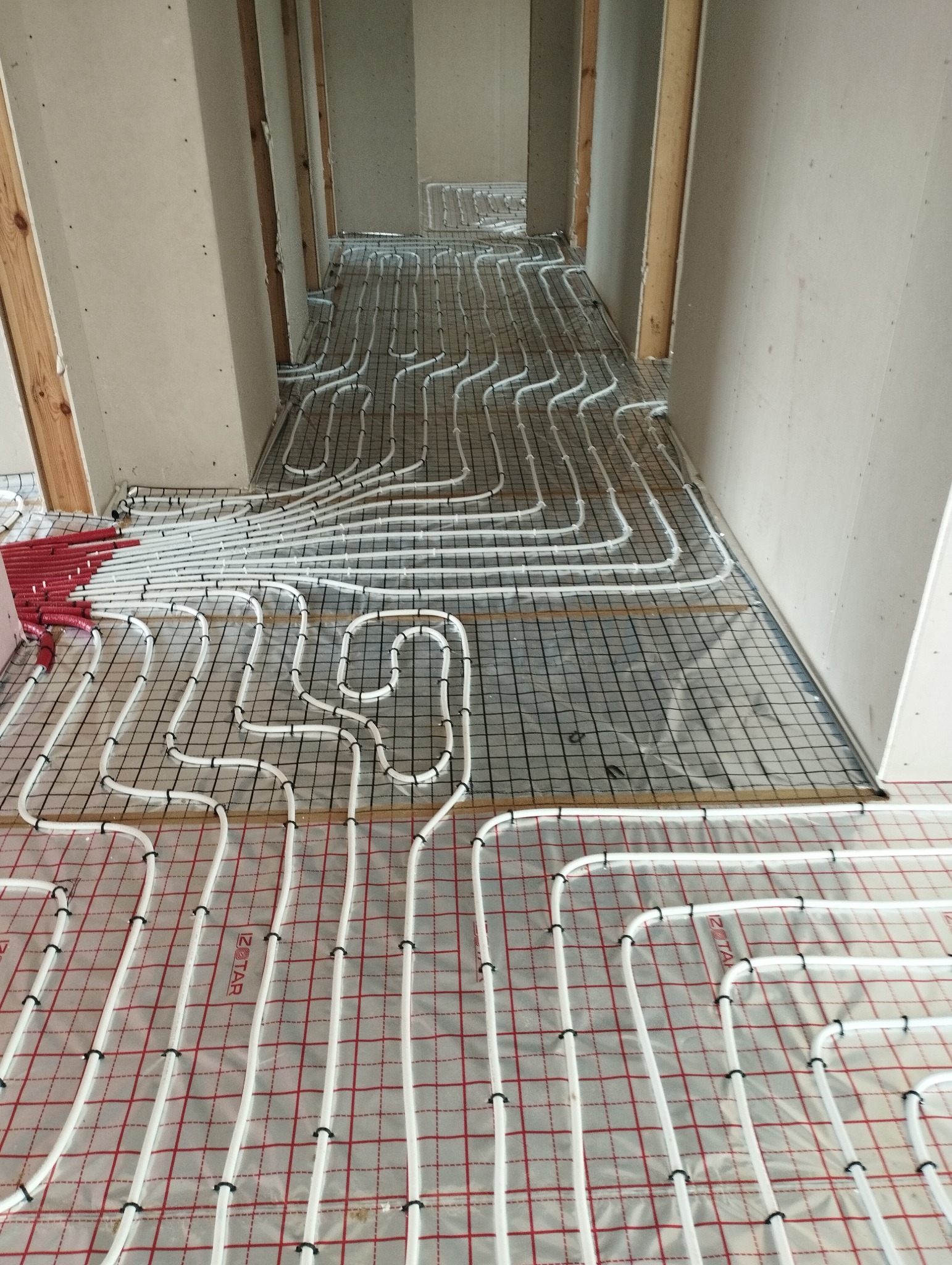 Białe rury ogrzewania podłogowego, przymocowane czarnymi klipsami do siatki, rozłożone na folii z czerwoną kratką, w surowym wnętrzu w trakcie instalacji.