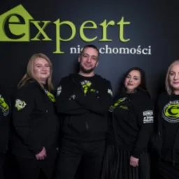 Zespół biura nieruchomości Expert w czarnych bluzach z logo firmy na tle ściany z nazwą firmy, mężczyzna w środku z założonymi rękami.