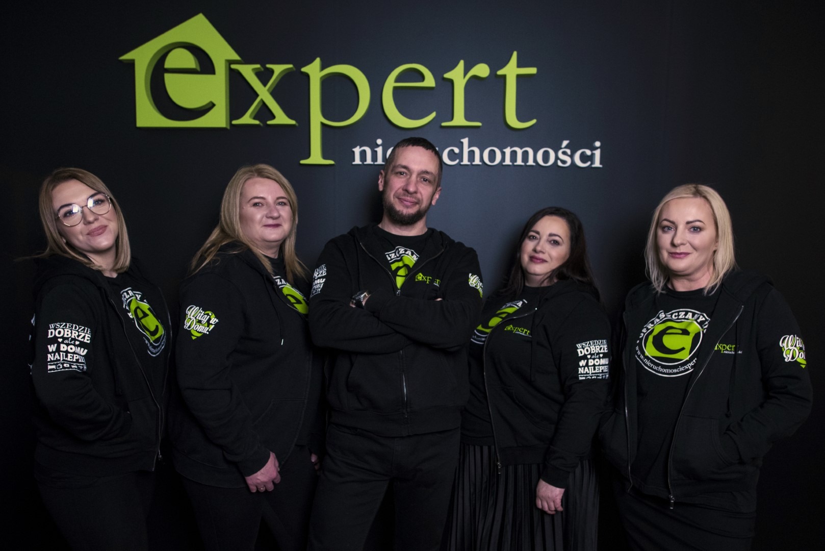 Zespół biura nieruchomości Expert w czarnych bluzach z logo firmy na tle ściany z nazwą firmy, mężczyzna w środku z założonymi rękami.