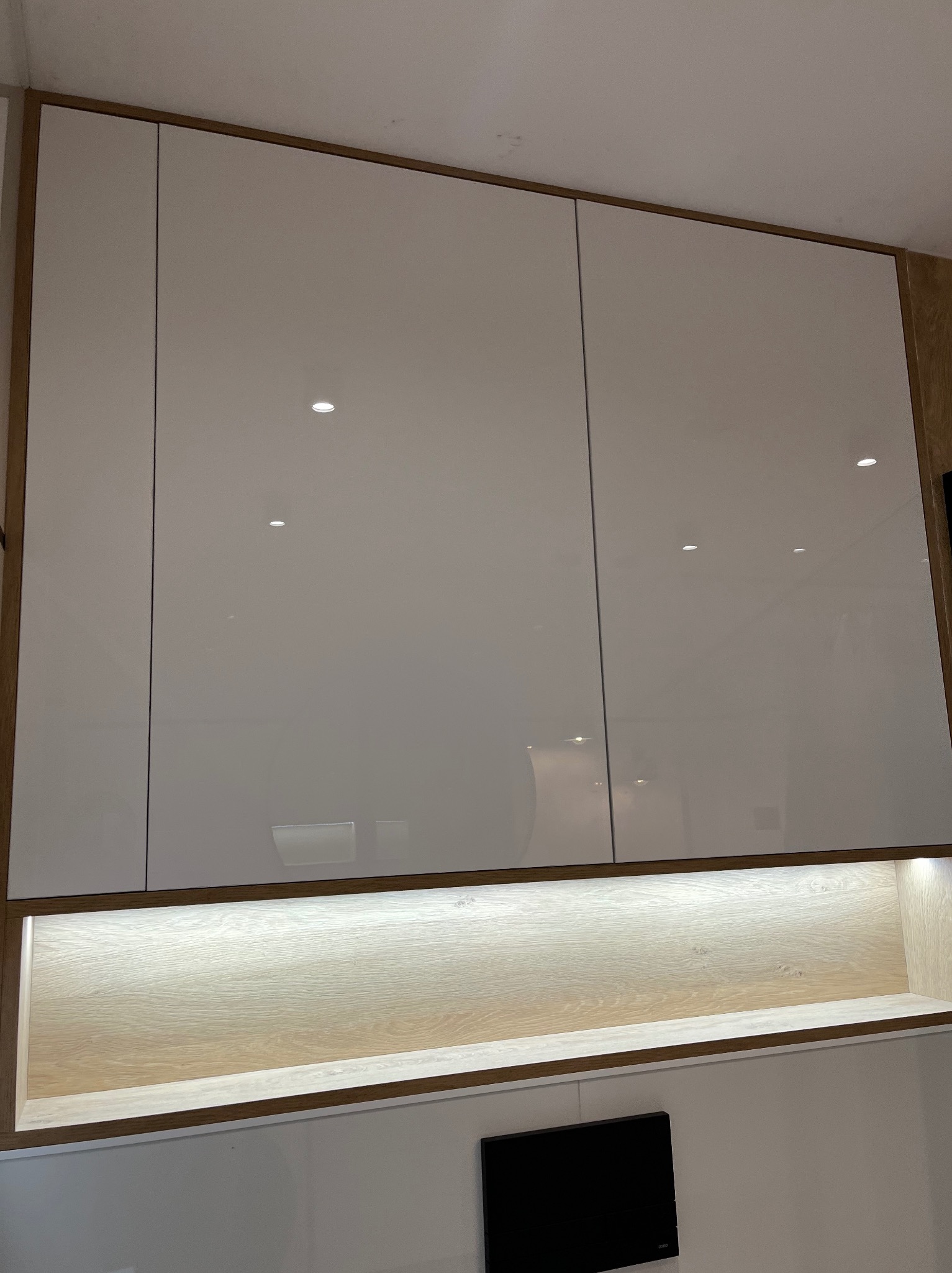 Białe, lakierowane szafki wiszące z drewnianym obramowaniem i oświetleniem LED, tworzące nowoczesny element zabudowy ściennej.