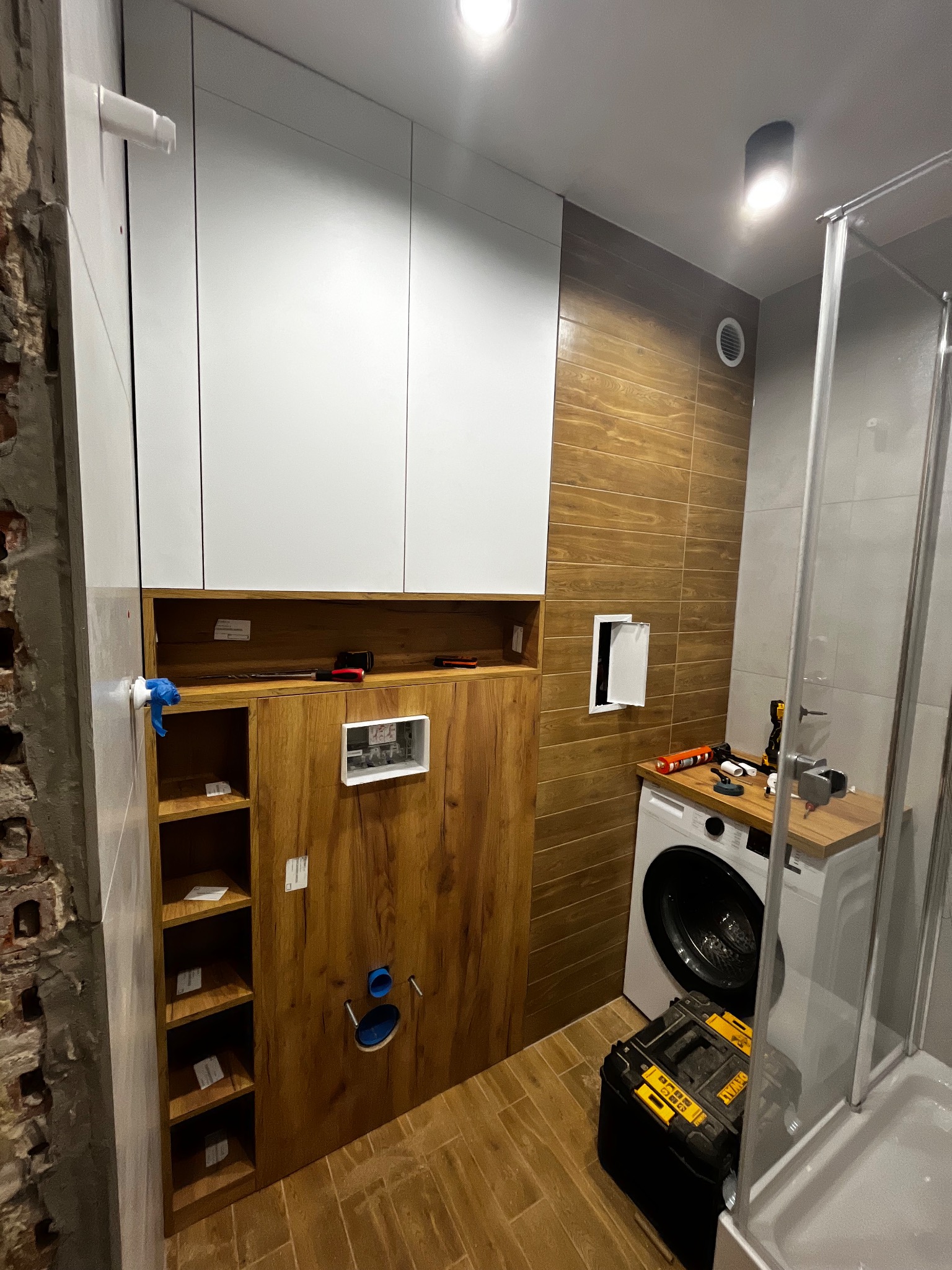 Montaż szafy wnękowej w łazience z drewnianym wykończeniem, widoczne narzędzia i pralka. Częściowo odsłonięta ściana z cegły dodaje surowego charakteru.