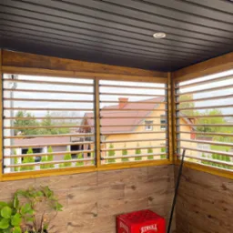 Zadaszony balkon z drewnianymi żaluzjami, widok na domy i zieleń, drewnopodobne płytki na ścianie, czerwona skrzynka Tyskie i roślina w doniczce w rogu.