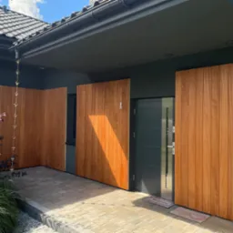 Nowoczesne wejście do domu z drewnianą zabudową, widok na drzwi i fragment elewacji. Ciekawy detal: łańcuch deszczowy.