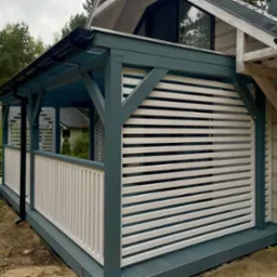 Zadaszona konstrukcja z białymi, poziomymi panelami i elementami w kolorze morskim. Widoczne drewniane słupy i fragment dachu budynku.