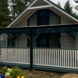 Drewniany domek letniskowy z zadaszonym tarasem i białą balustradą. Elementy konstrukcyjne w kolorze ciemnej zieleni. W tle widoczne drzewa.