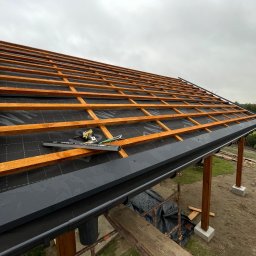 Easy Finish Michał Walczak - Konstrukcja dachu altany z drewnianymi belkami i czarną membraną, widoczne narzędzia i elementy mocujące, w tle pochmurne niebo. Etap budowy z bliska.