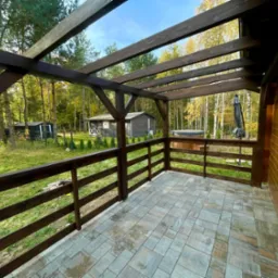 Zadaszony taras z drewnianą konstrukcją i balustradą, wyłożony kostką brukową. W tle widoczne budynki gospodarcze i drzewa. Ujęcie z perspektywy narożnika.