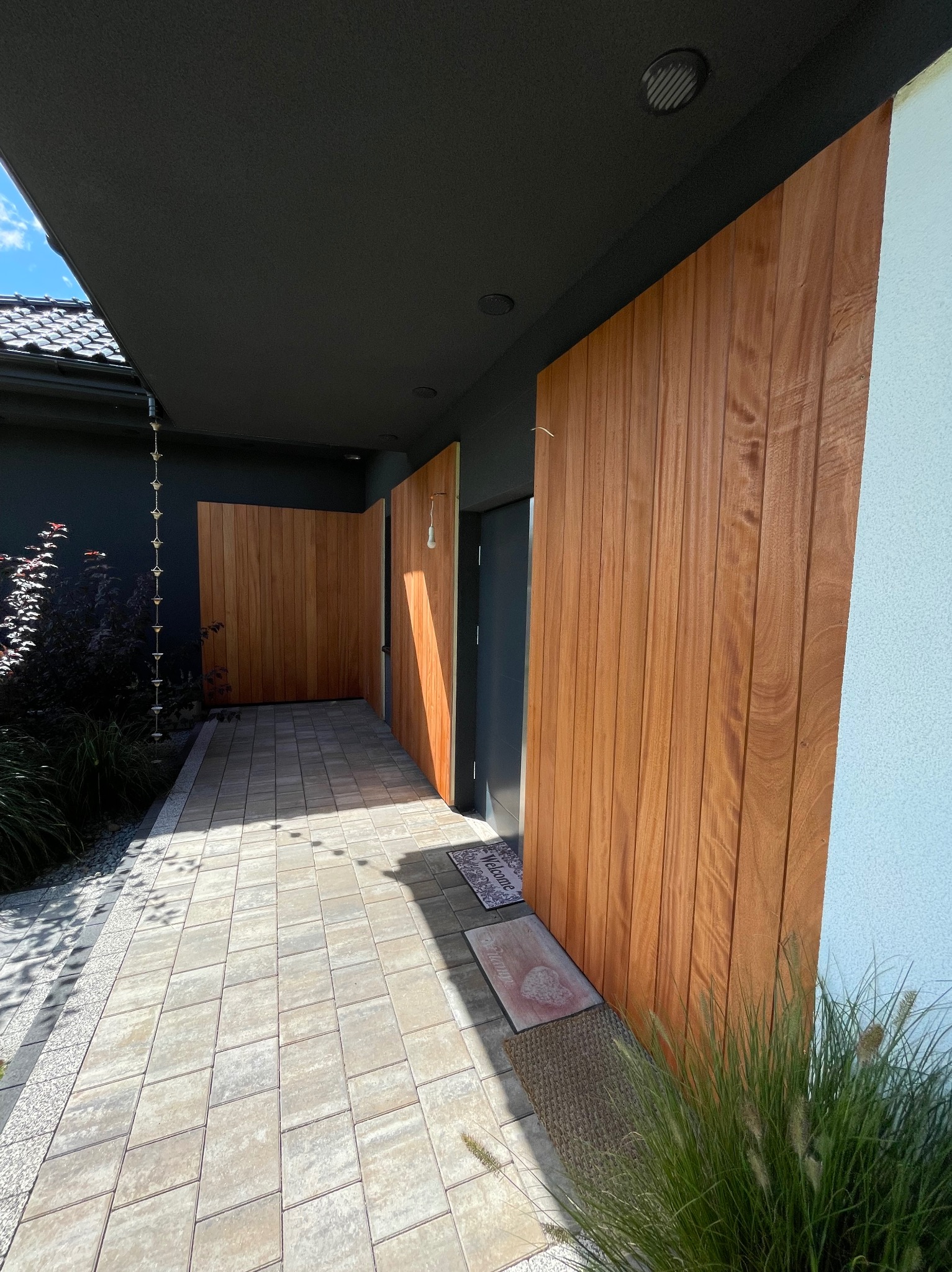 Nowoczesne wejście do domu z pionową okładziną z drewna o ciepłym odcieniu, kontrastującą z ciemnym sufitem podcienia i jasną elewacją. Widoczna kostka brukowa i dekoracyjna roślinność.