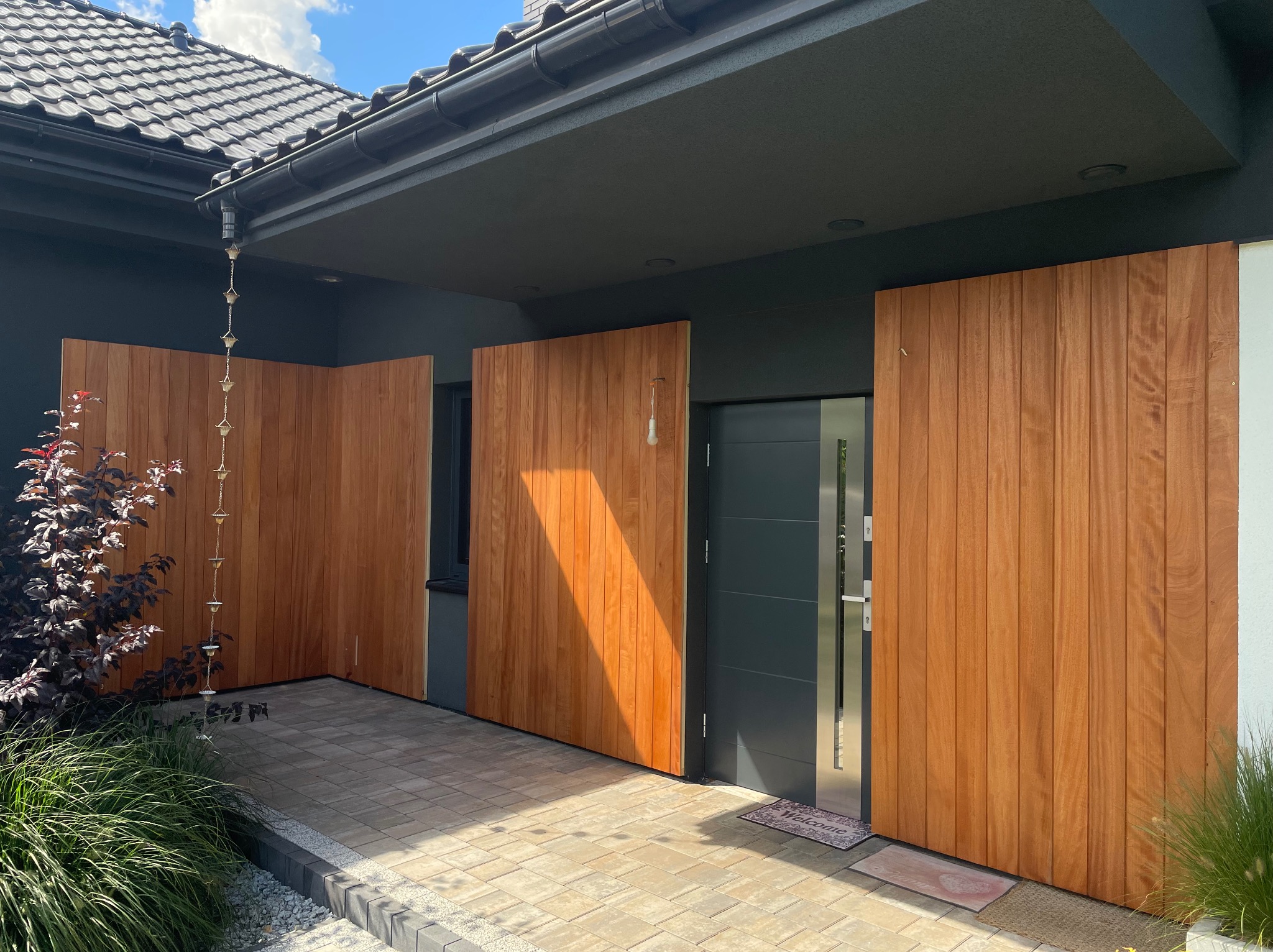 Nowoczesne wejście do domu z drewnianą zabudową, widok na drzwi i fragment elewacji. Ciekawy detal: łańcuch deszczowy.